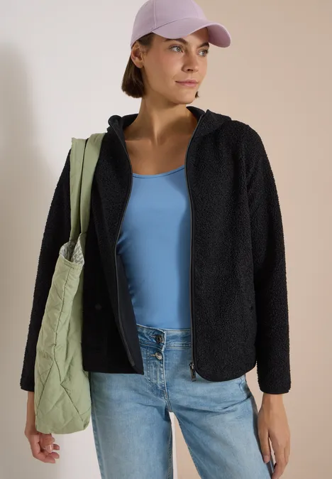 Frau mit Baseball-Cap, hellblauem Top, schwarzer Jacke, Jeans und grünem Stepp-Shopper blickt seitlich.