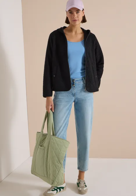 Frau im Casual-Look mit schwarzer Jacke, blauem Top, Jeans, Sneakers und grünem Shopper.