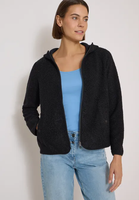 Frau trägt schwarze Teddy-Fleecejacke mit Kapuze, hellblaues Top und Jeans vor weißem Hintergrund.