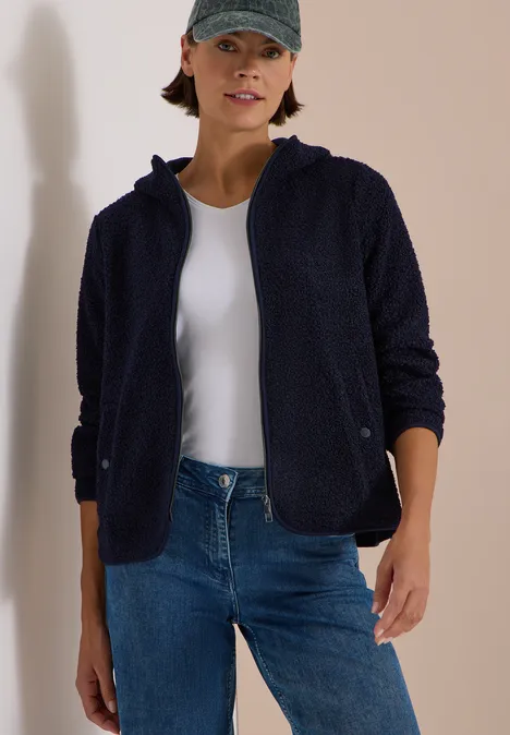 Frau in dunkelblauer Fleecejacke mit Kapuze, weißem Shirt, Jeans und Baseballcap vor hellem Hintergrund.