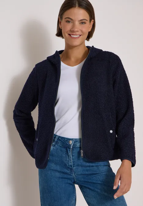Lächelnde Frau in dunkelblauer Fleecejacke, weißem T-Shirt und Jeans vor hellem Hintergrund.