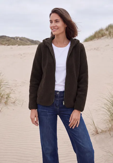 Teddy-Jacke mit Kapuze in Unifarbe truffle brown Frau in dunkelgrüner Fleecejacke, weißem T-Shirt und blauen Jeans steht lächelnd am Dünenstrand