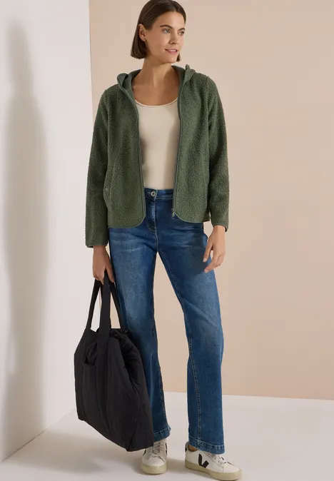 Frau in grüner Jacke, beigem Top, Jeans und weißen Sneakern, sie hält eine schwarze Tasche.