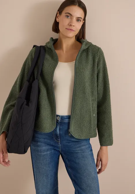 Frau in grüner Fleecejacke, weißem Top, Jeans und schwarzer gesteppter Tasche vor beigem Hintergrund.