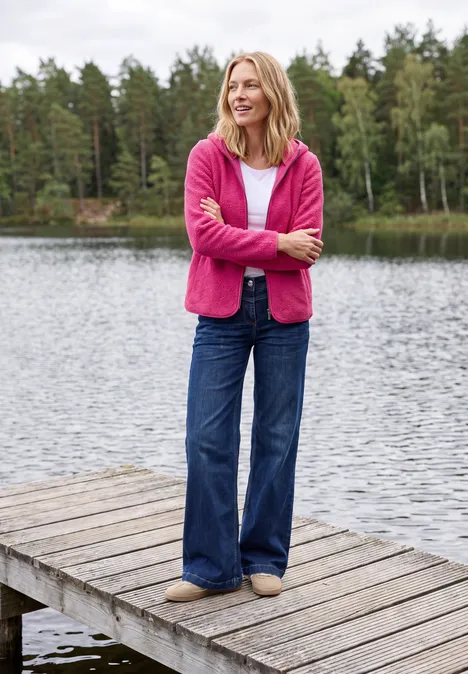 Eine Frau in pinkem Pullover und Jeans steht entspannt auf einem Holzsteg am See, Wald im Hintergrund.