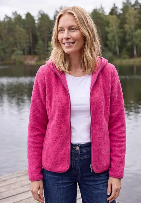 Frau steht am Seeufer auf einem Steg, trägt pinke Fleecejacke, weiße Bluse und Jeans.