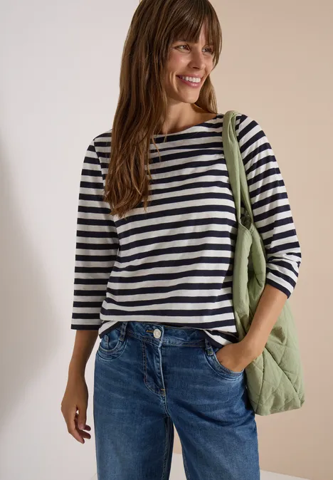 Frau mit gestreiftem 3/4-Arm-Shirt, Jeans und grüner Stepp-Umhängetasche lächelt vor neutralem Hintergrund.