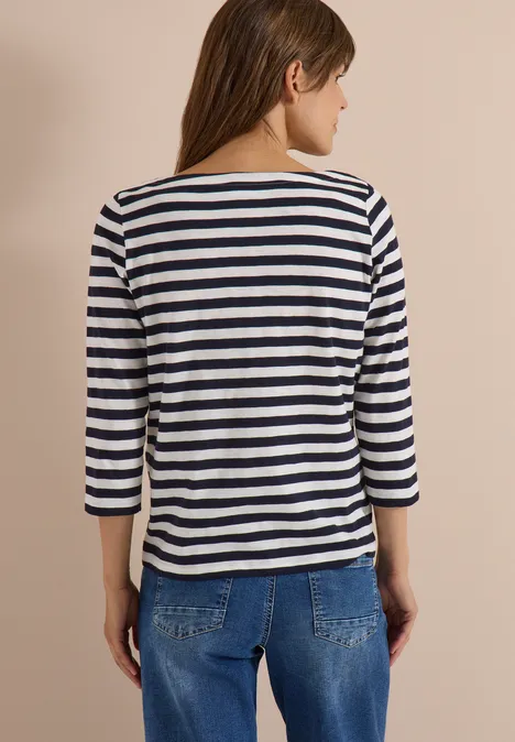 Rückansicht einer Frau mit gestreiftem Dreiviertelarmshirt und blauen Jeans vor hellem Hintergrund.