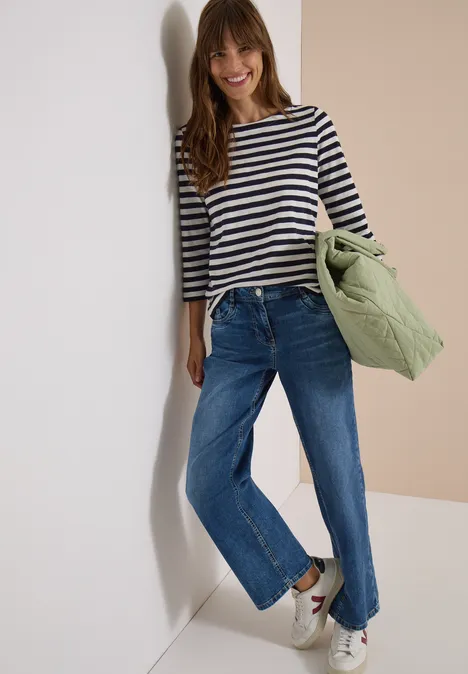 Eine lächelnde Frau in gestreiftem Shirt, blauer Jeans und Sneakers hält eine grüne Steppjacke vor hellem Hintergrund.