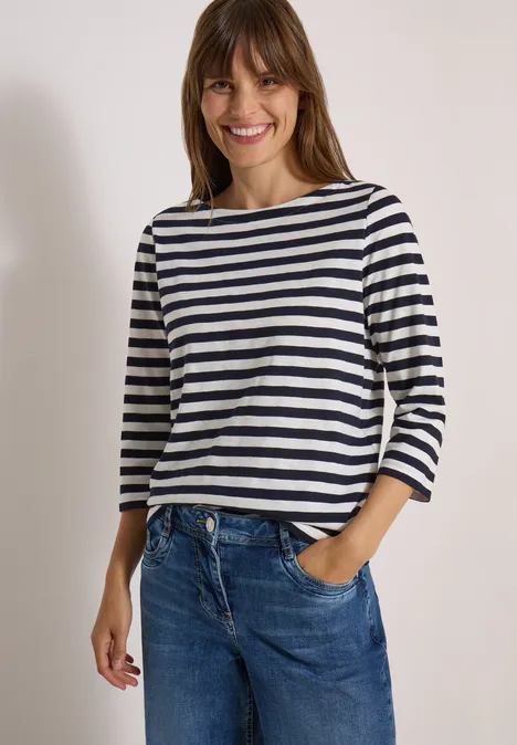 Junge Frau lächelt in blau-weiß gestreiftem Dreiviertelarmshirt und blauen Jeans vor schlichtem Hintergrund.
