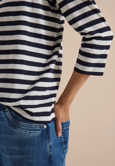 Oberkörper in weiß-blau gestreiftem Shirt, Hand in Jeans-Tasche vor beigem Hintergrund.