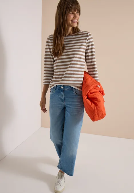 T-shirt basique à col bateau et rayures natural taupe Frau in gestreiftem Pullover, weiten Jeans und Sneakern hält eine orange Jacke und lächelt.