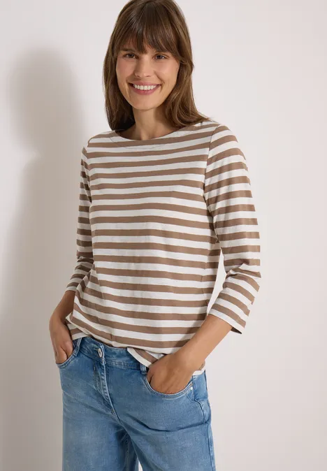 T-shirt basique à col bateau et rayures natural taupe Fröhliche junge Frau mit braun-weißer Ringelshirt und blauen Jeans vor heller Wand.