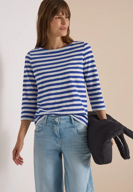 Frau trägt blau-weiß gestreiftes Langarmshirt, helle Jeans und hält eine dunkle Jacke locker in der Hand.