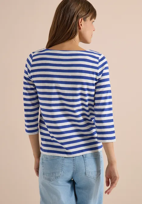 Eine Frau von hinten in blau-weiß geringeltem 3/4-Arm-Shirt und hellen Jeans vor beigem Hintergrund.
