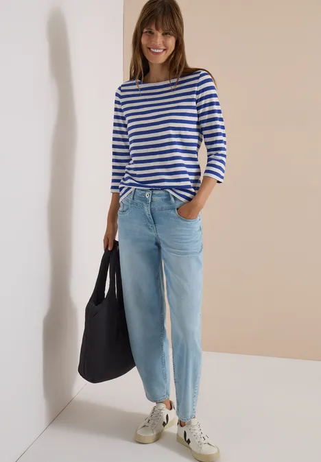 Frau in blau-weiß gestreiftem Pullover, hellen Jeans, Sneakers und schwarzer Tasche vor beigem Hintergrund.