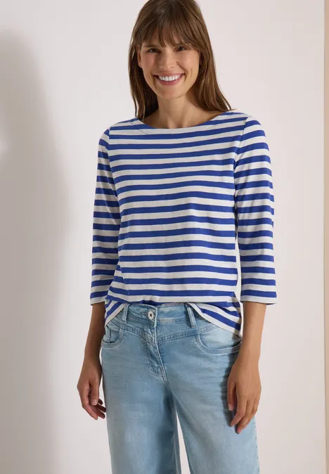 Frau trägt blau-weiß gestreiftes 3/4-Arm-Shirt und helle Jeans, lächelt.