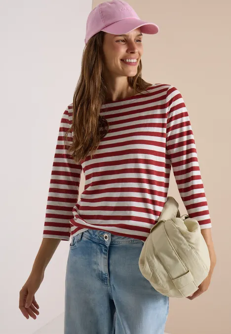 Frau in rot-weiß gestreiftem Shirt, Jeans und rosa Baseballcap trägt lässige Tasche und lächelt.