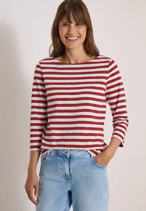 Eine lächelnde Frau trägt ein rot-weiß gestreiftes Shirt und Jeans vor neutralem Hintergrund.