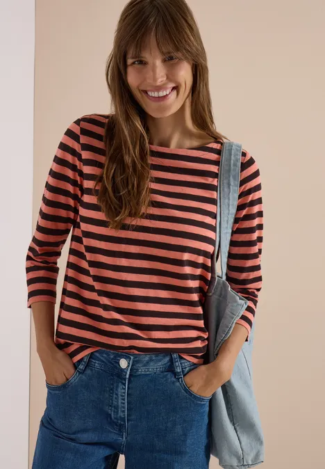 Frau im gestreiften 3/4-Arm-Shirt und Jeans, lächelnd mit Händen in den Taschen vor beigem Hintergrund.