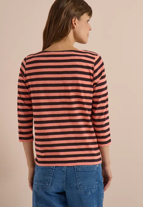 Frau von hinten in rosa-schwarz gestreiftem 3/4-Arm-Shirt und blauen Jeans vor beiger Wand.
