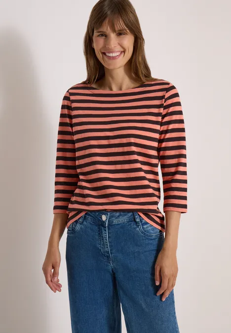 Lächelnde Frau in rotschwarz gestreiftem Shirt und blauer Jeanshose vor hellem Hintergrund.