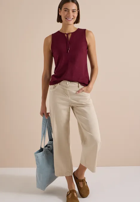 Kobieta w beżowych culottes, bordo topie bez rękawów, brązowych mokasynach i niebieskiej torbie.
