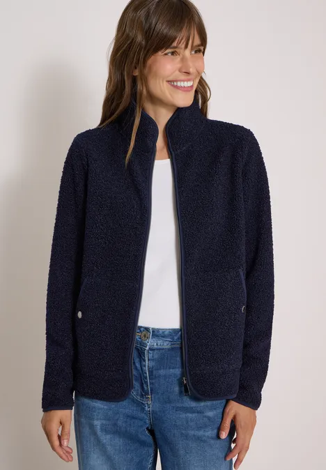 Frau trägt dunkelblaue Fleecejacke und Jeans, lächelt vor neutralem Hintergrund.