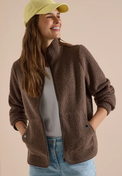 Lächelnde Frau in brauner Teddyfleece-Jacke, grauem Shirt und hellgrüner Cap.