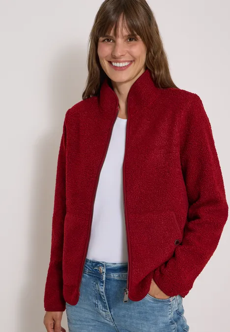 Eine lächelnde Frau trägt eine rote Fleecejacke mit Reißverschluss und blaue Jeans vor weißem Hintergrund.