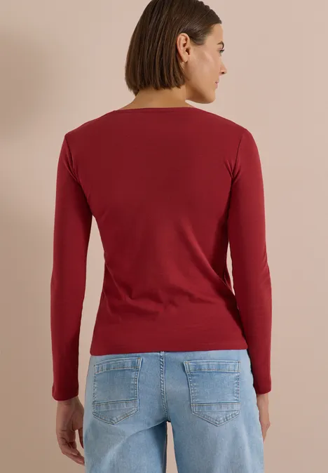 Rückansicht einer Frau in rotem Langarmshirt und hellblauen Jeans vor hellem Hintergrund.