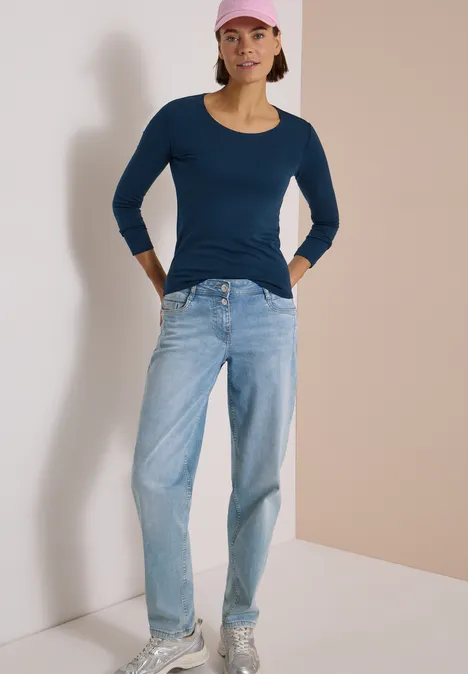 Frau in Jeans, blauem Longsleeve und rosa Cap lehnt entspannt an einer Wand.