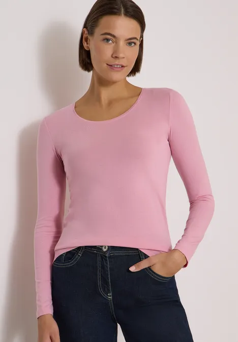 Frau in rosa langärmeligem Oberteil und dunklen Jeans steht lächelnd mit einer Hand in der Tasche.