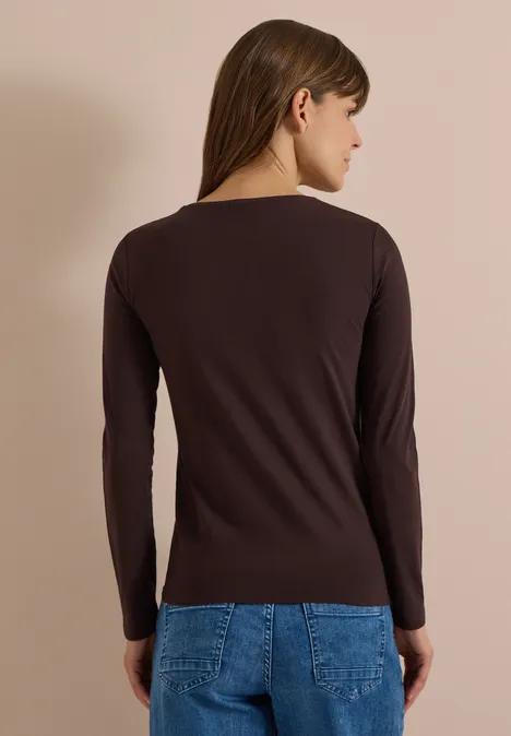 Rückansicht einer Frau in braunem Langarmshirt und blauer Jeans vor beigem Hintergrund.
