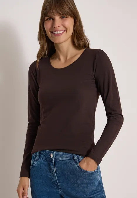 Frau in dunklem Langarmshirt und Jeans, lächelt, steht mit einer Hand in der Hosentasche vor neutralem Hintergrund