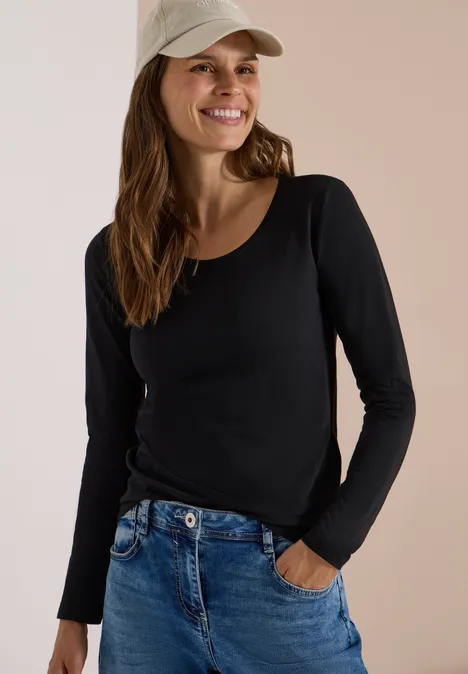 Basic Shirt mit Rundhals im 2er-Pack cashmere beige/black Eine lächelnde Frau in schwarzem Langarmshirt, blauen Jeans und beiger Basecap, Hand in Tasche.