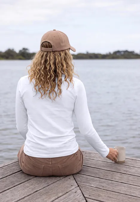 Une femme aux cheveux bouclés et casquette est assise sur un ponton, tient une tasse et regarde le lac.