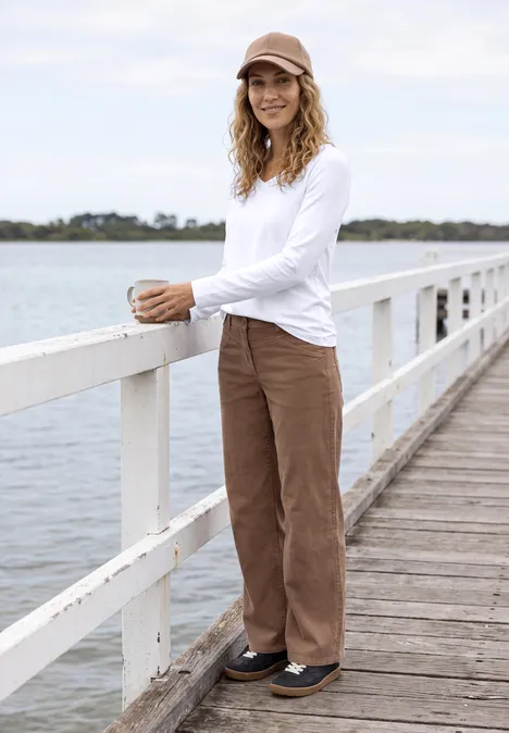 Femme en haut blanc, pantalon marron et casquette, posant sur une jetée au bord de l'eau.