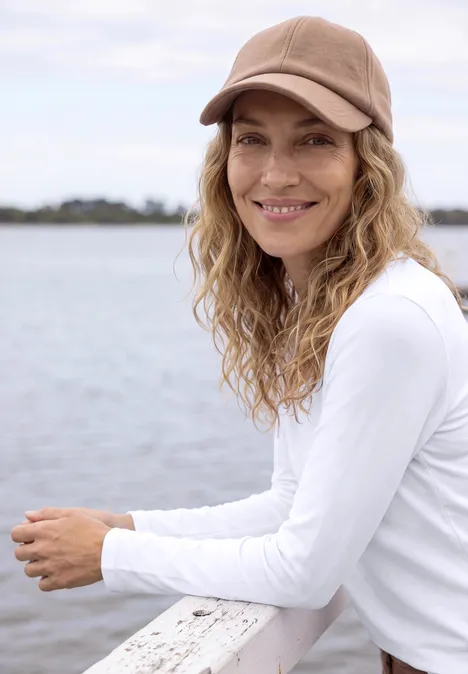 Une femme souriante en casquette beige et tee-shirt blanc appuyée sur une rambarde au bord de l'eau.