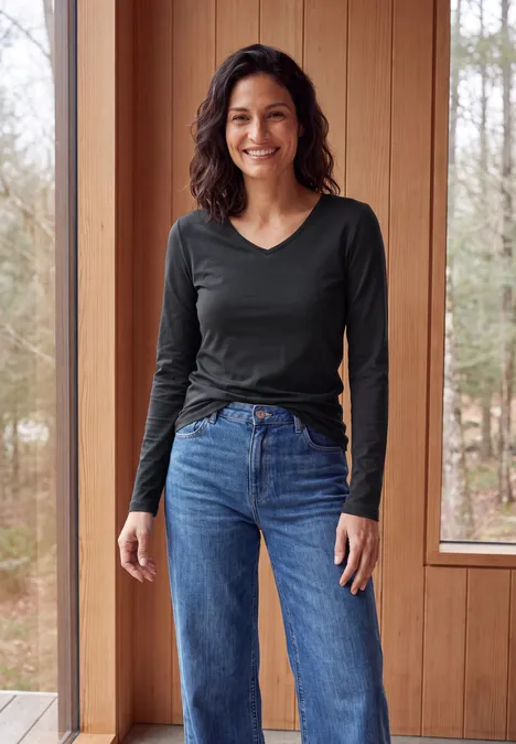 Femme souriante aux cheveux foncés, t-shirt noir à manches longues et jean, debout devant une fenêtre en bois.