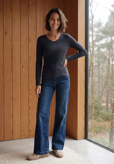Longsleeve met V-hals urban dark blue Eine lächelnde Frau in langärmeligem Shirt und weiten Jeans steht vor einer Holz-Wand und Fenster mit Waldblick.
