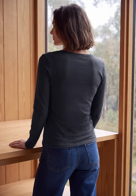 Longsleeve met V-hals urban dark blue Eine Frau mit dunklen Haaren steht an einem Fenster in einem Holzrahmen, trägt Jeans und langärmeliges Shirt.