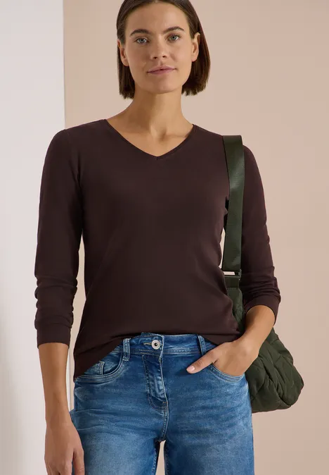 Frau in braunem V-Pullover, blauer Jeans, mit Hand in Tasche und grüner Umhängetasche.