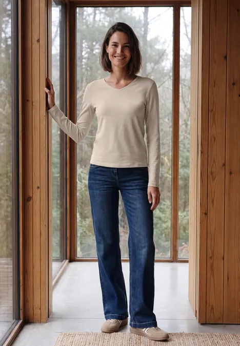 Longsleeve met V-hals cashmere beige Eine lächelnde Frau in hellem Pullover und Jeans steht vor bodentiefen Holzfenstern.