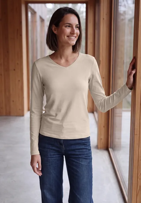 Longsleeve met V-hals cashmere beige Lächelnde Frau im beigen Langarmshirt und Jeans lehnt an Fensterrahmen in modernem Holzinterieur.