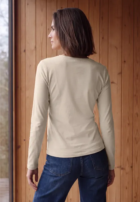 Longsleeve met V-hals cashmere beige Frau in langärmeligem beigem Shirt und Jeans, Rückansicht vor einer Holzverkleidung.