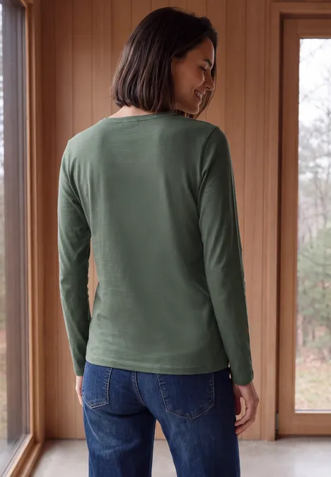 Langarm Shirt in Unifarbe mit V-Neck dried olive green Frau steht von hinten in grünem Langarmshirt und Jeans vor großen Fenstern.