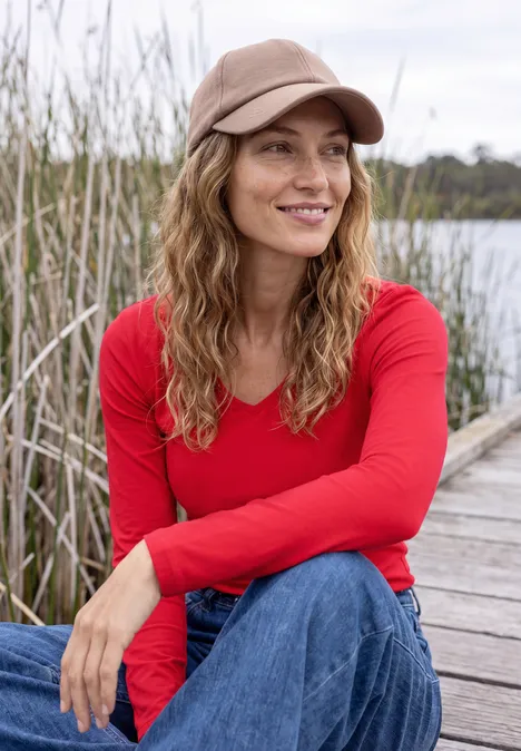 Une femme en t-shirt rouge et casquette est assise sur une jetée au bord d’un lac, entourée de roseaux.
