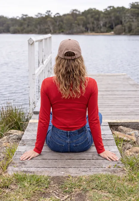 Une personne en pull rouge et casquette est assise sur un ponton, contemplant un lac tranquille.