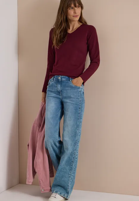 Langarm Shirt in Unifarbe mit V-Neck red wine Frau in weiten Jeans und burgunderfarbenem Longsleeve, hält rosa Jacke, vor schlichtem beigem Hintergrund.